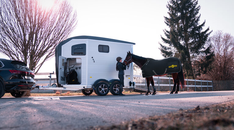 A woman is standing next to her horse, stroking it. In the background, you can see the Equitos Alu, which is coupled to a vehicle.