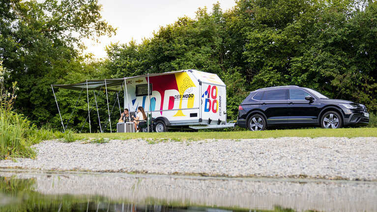 Remorque multifonctionnelle MODO avec accessoires de camping se trouve sur une prairie au bord d'un lac. | © Humbaur GmbH
