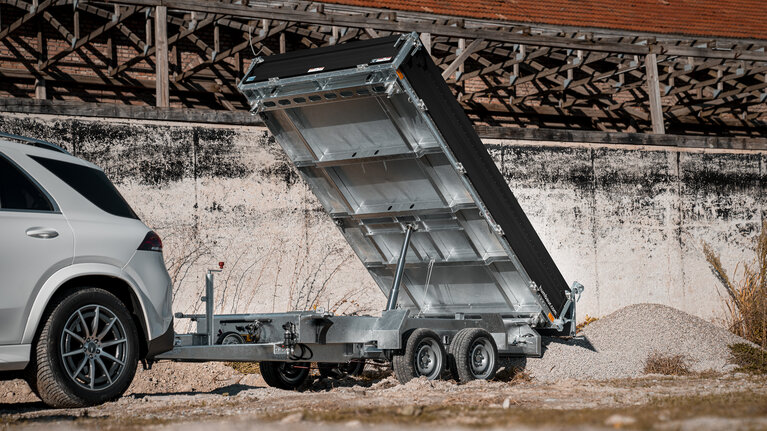 Construction material is tipped backwards from a three-way tipper. The trailer is coupled to a car.  | © Humbaur GmbH